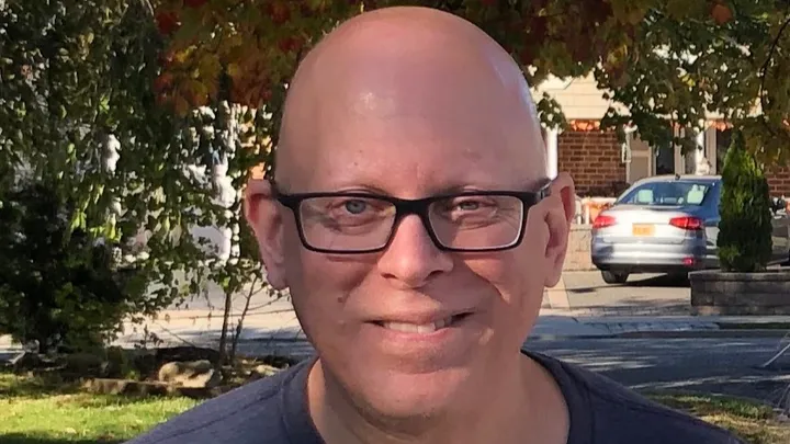 Shallel Inglima smiling warmly outdoors, wearing dark-framed glasses